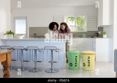 Afroamerikanische Mutter und jugendliche Tochter lehnen sich an der Insel und betrachten das Telefon, während sie eine Keramik-Tasse halten Stockfoto