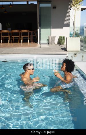 Afroamerikanische Männer, die auf dem Rand eines Dachpools in Badehosen sitzen und Cocktails halten, mit Sonnenbrillen. Duo, lässig, im Freien, Terrasse Stockfoto