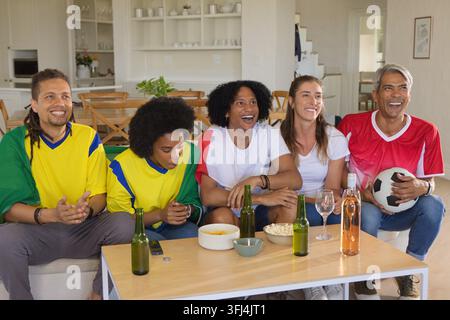 Verschiedene Freunde in Fußballtrikots, die ein Spiel im Wohnzimmer mit Bierflaschen und einem Fußball ansehen. Sport, Gruppe, Zuhause, lässig, Lounge, Fernsehen, Stockfoto