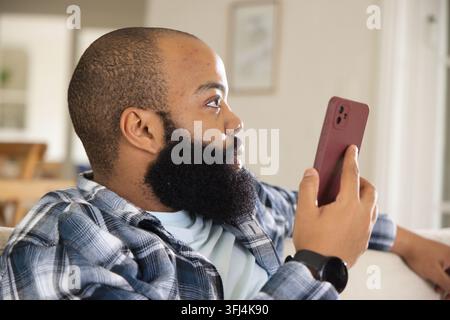 Erwachsener Mann sitzt zu Hause auf der Couch, hält ein Smartphone mit brauner Hülle und trägt ein kariertes Flanellhemd. Gerät, Fokus, Konnektivität, Stockfoto