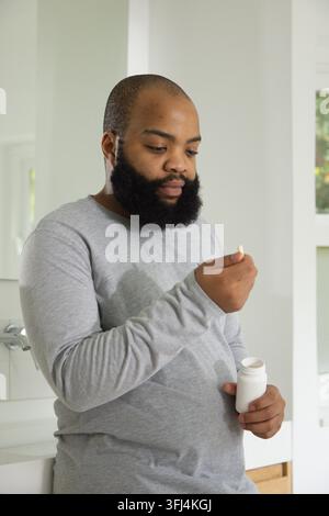 Afroamerikaner hält eine weiße Pille und ein Pillenfläschchen, während er sein feuchtes Hemd am Waschbecken im Badezimmer überprüft. Bart, Waschraum, Medikamente, Stockfoto
