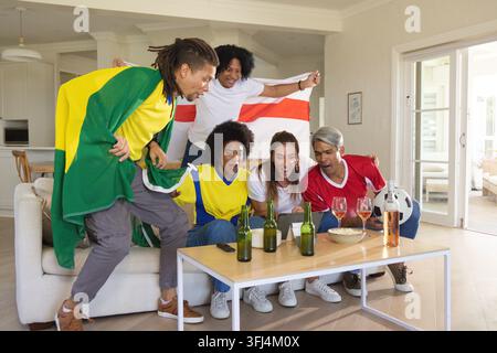 Verschiedene Freunde in Trikots, die auf einem Sofa mit Bierflaschen und einem Fußball auf ein Tablet jubeln und schauen. Fans, Gruppe, Betrachtung, Bildschirm, Flagge, Teamfarben, Livestream Stockfoto