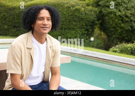 Afroamerikaner sitzt am Poolrand im Garten, trägt ein beiges Hemd, Ohrring und Armband, Kopierraum. Hinterhof, Wasser, Terrasse, Hecke, Rasen, Garten, draußen Stockfoto