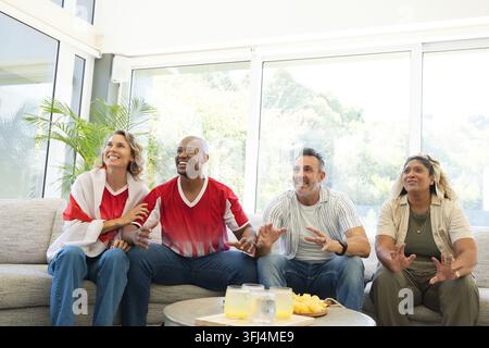 Verschiedene Freunde jubeln auf grauem Sofa zu Hause am Kaffeetisch, zwei tragen rote Sportoberteile. Treffen, Zuschauen, sonnenbeschienen, Wohnzimmer, modern, Interieur Stockfoto