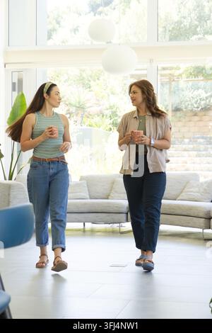 Zwei Frauen gehen nebeneinander in einem hellen Wohnzimmer, halten Keramikbecher und unterhalten sich. Freunde, Gespräch, Zuhause, gemütlich, sonnenbeschienen, Fenster, Pflanzen Stockfoto