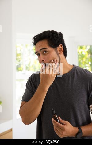 Afroamerikanischer Mann trägt Serum aus einer Bernsteinflasche auf dem heimischen Schminktisch auf, trägt eine schwarze Uhr. Hautpflege, Selbstpflege, Wellness, Tageslicht, Badezimmer, Grünpflanzen, Stockfoto
