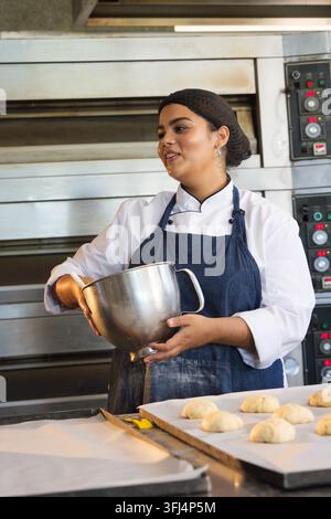 Afrikanische Frau trägt Chefjacke und hält eine Edelstahl-Rührschüssel an einer Backbank und formt Teig. Bäcker, Küche, Öfen, Decköfen, Brot, Backpapier Stockfoto