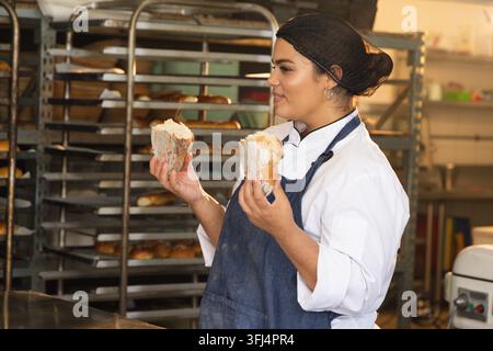 Afroamerikanische Bäckerin in Chefjacke, Schürze inspiziert zwei Brotlaibhälften in der Bäckerei, Kopierraum. Brot, Ausrüstung, Produktion, industriell, metallisch Stockfoto