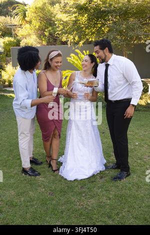Verschiedene Gruppe, die auf dem Gartenrasen anstößt, Braut in weißem Kleid, Bräutigam, der Champagner in Sektgläser eingießt. Hochzeit, Paar, Hinterhof, draußen, Feier, Anzug Stockfoto
