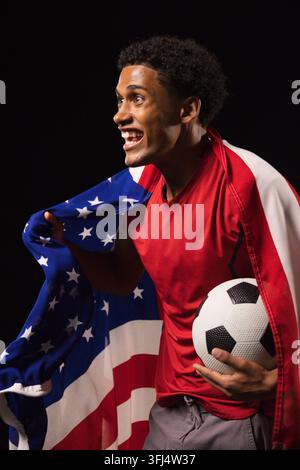 Afroamerikaner in rotem Hemd, der eine US-Flagge drapiert, hält im Studio einen Fußball. Athlet, Werbung, Kampagne, dramatisch, hochkontrast Stockfoto