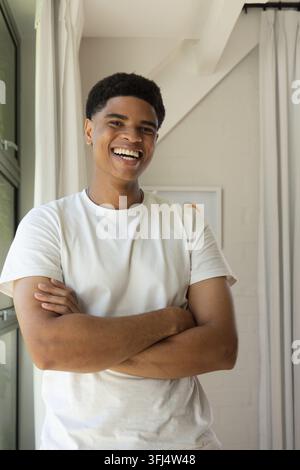 Afroamerikanischer Mann steht im Studio an einem großen Fenster, lächelt mit verschränkten Armen und Kunst im Hintergrund. Porträt, Solo, Ziegelwand, Vorhänge, Druck, natürlich Stockfoto