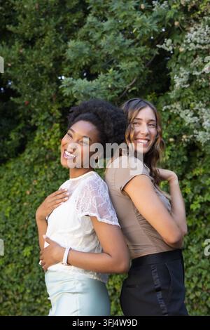 Verschiedene Freundinnen, die Rücken an Rücken posieren und in den Garten lächeln, mit Kreolen geschmückt. Draußen, Duo, Laub, Blumen, lässig, natürlich, modisch Stockfoto