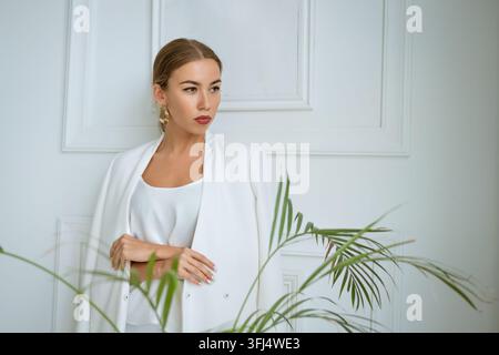 Kaukasische Geschäftsfrau in einem weißen Blazer steht mit verschränkten Armen und seitwärts gewandtem Blick vor einer vertäfelten Wand, ein grünes Palmblatt in der Vor… Stockfoto