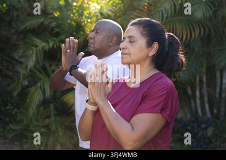 Afroamerikanisches Paar meditiert in sonnigem Hof mit Palmen, trägt magenta Oberteil, weißes T-Shirt, Uhren. Hinterhof, tropisch, Grün Stockfoto