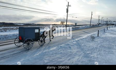 Amish-Buggy auf verschneiter Straße in Lancaster County, PA, unter einem Sonnenuntergangshimmel. Luftaufnahmeschwenk, während der Schnee auf ländlicher USA-Straße liegt, Strasburg, Pennsylvania. Stockfoto