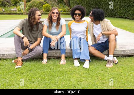 Verschiedene Freunde sitzen auf dem Rasen an einem hölzernen Pooldeck, tragen lässige Kleidung und Sonnenbrillen. Gruppe, draußen, Hinterhof, Rasen, Garten, gestaltet, rela Stockfoto