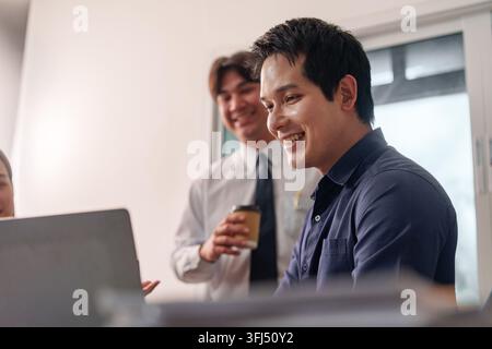 Zwei glückliche asiatische Geschäftsleute diskutieren Projektdetails vor einem Laptop-Bildschirm in einer hellen modernen Büroumgebung. Stockfoto