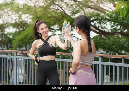Zwei fitte asiatische Frauen in Sportkleidung halten klare Wasserflaschen hoch, um nach dem Training im Park anzustoßen. Stockfoto