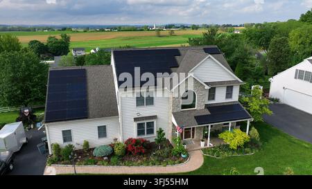 Amerikanisches Einfamilienhaus, das grüne Energie mit Solarpaneelen auf dem Dach produziert. Winkende amerikanische Flagge am Eingang. Ländliche Farmfelder in der Ferne. Luftbild Stockfoto