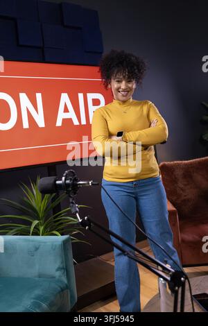 Stehende, afroamerikanische Frau in einem Senfgelben Oberteil im Studio in der Nähe eines ON AIR-Schildes und eines Mikrofons Stockfoto