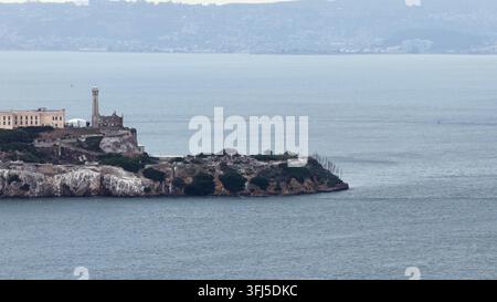 Der historische Leuchtturm und die verlassenen Gefängnisgebäude von Alcatraz Island erheben sich über felsige Klippen, trübe Gewässer der San Francisco Bay erstrecken sich in Richtung entfernter Hügel Stockfoto