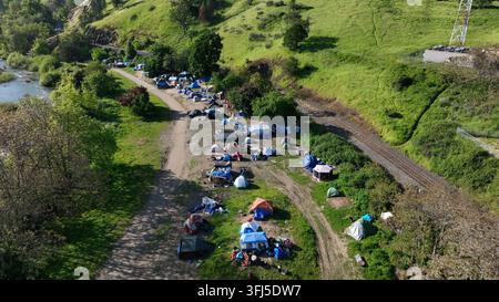 Obdachlosenlager mit bunten Zelten und blauen Planen breitet sich entlang von Schotterwegen neben dem Fluss unter einem üppigen grünen Hügel an einem sonnigen Frühlingstag in Roseb aus Stockfoto