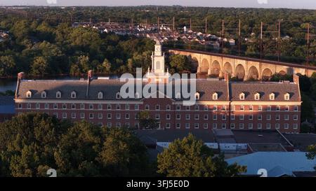 Historischer Uhrturm krönt das Gebäude der Rutgers University bei goldener Sonnenaufgang in New Brunswick, New Jersey, steinerner Eisenbahnviadukt spannt sich über den Fluss dahinter Stockfoto