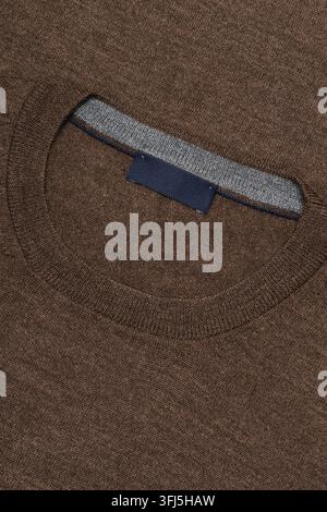 Nahaufnahme eines braunen gestrickten Kleidungsausschnitts mit grauer Bordüre und einem leeren dunkelblauen Stoffetikett. Die detaillierte Textur und die gerippte Konstruktion sind Stockfoto