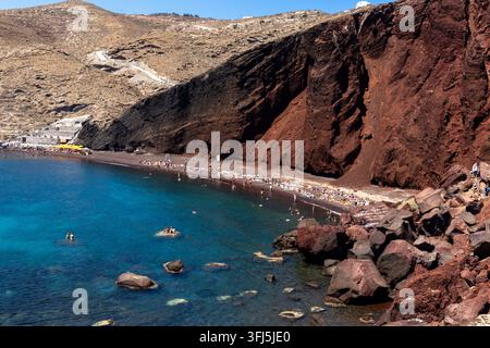 Panoramablick auf Red Beach (Kokkini Paralia) in Santorini, Griechenland, mit dramatischen roten Vulkanfelsen, dunklem Sand, türkisfarbenem Wasser und Touristen e Stockfoto