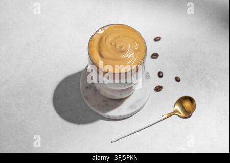 Dalgona-Kaffee. Geschlagener Instant-Eiskaffee in einem Glas mit Kaffeebohnen, Löffel und Schatten auf einem grauen Betonhintergrund. Trendiges süßes Dessertkonzept Stockfoto