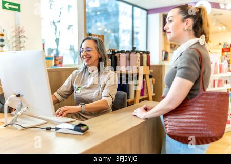 Apotheker mit Brille arbeitet am Computer und lächelt fröhlich, während er einer weiblichen Kundin hilft, die am Tresen in einer modernen Einzelhandelsapotheke steht Stockfoto