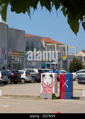 Pirovac, Kroatien, 16. August 2026 Urbaner Meeresufer-Parkplatz mit niedrigen Betongebäuden, geparkten Autos, Ladenfronten, Touristenbüro und strahlend blauem Himmel Stockfoto