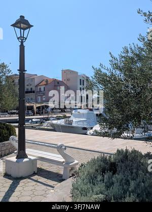 Pirovac, Kroatien, 16. August 2026 Ruhige Uferpromenade neben festgemachten Booten, pastellfarbene Dorfgebäude, Olivenbäume und klarer blauer Himmel, die Stockfoto