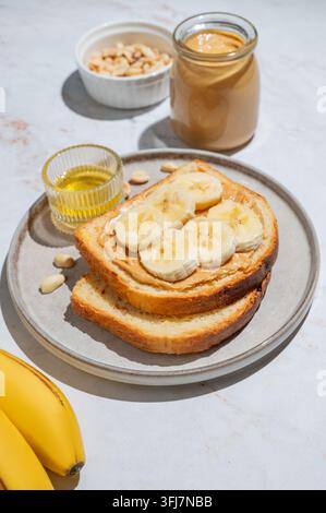 Erdnussbutter-Toast und Banane auf einem Teller auf einem Marmorhintergrund mit Früchten, Nüssen, Honig und Morgenschatten. Konzept von süßer Ernährung und gesunden Snacks Stockfoto