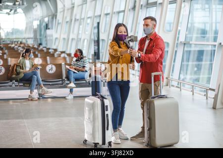 Ein Mann und eine Frau mit Gesichtsbedeckungen nutzen einen Smartphone-Gimbal, um ein Video aufzunehmen, während sie auf ihren Flug an einem modernen Flughafen warten Stockfoto