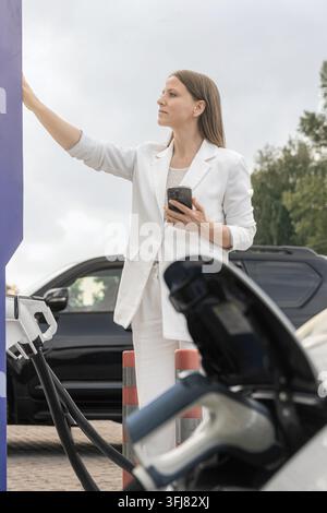 Junge Frau, etwa 25–35 Jahre alt, in einem weißen Anzug, die sich nach dem Ladestation-Panel streckt, während sie in der anderen Hand ein Smartphone hält. Die Stockfoto