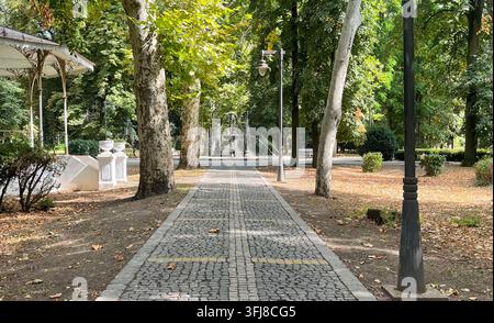 Ein ruhiger gepflasterter Weg führt durch einen grünen Garten mit alten Bäumen, Parklaternen und gefallenen Blättern, die im sanften Herbstsonnenlicht über den Boden verstreut sind. Stockfoto