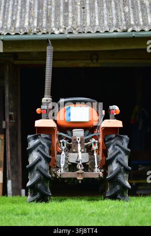 Rückansicht eines kompakten orangefarbenen Traktors, der vor einer rustikalen Holzbarn geparkt ist. Traditionelle landwirtschaftliche Maschinen und authentische Bauernhoflandschaft Stockfoto