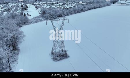 Luftbild-Drohnenaufnahme eines Hochspannungsübertragungsturms, der in einem schneebedeckten Feld in der Nähe einer Wohngegend im Winter in den Vereinigten Staaten steht Stockfoto