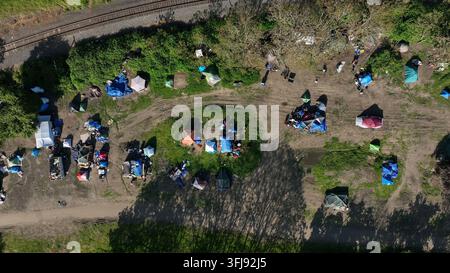 Ansicht von oben über ein weitläufiges Obdachlosenlager mit verstreuten Zelten, blauen Planen und Trümmern entlang abgenutzter Erdwege neben aktiven Eisenbahngleisen in Ro Stockfoto