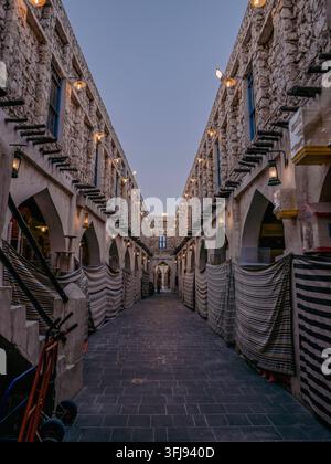 Symmetrische Niederwinkelaufnahme einer schmalen historischen Stein-Gasse mit beleuchteten Laternen und überdachten Marktständen im Souq Waqif. Stockfoto