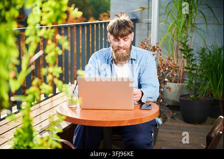 Lächelnder Freiberufler, der einen Notebook-Computer auf einem Balkoncafé benutzt, sitzt im warmen Tageslicht mit Getränk, Mobiltelefon, Geländer und Grünpflanzen am Tisch Stockfoto