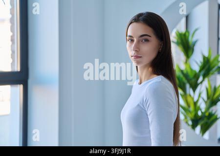 Porträt einer ernsten jungen Frau, die neben dem Fenster steht, seitliche Ansicht Stockfoto