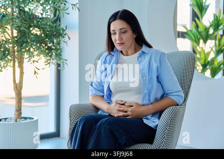 Mittlere Frau, die ihren Bauch hält und unter Bauchschmerzen in einem Sessel leidet Stockfoto
