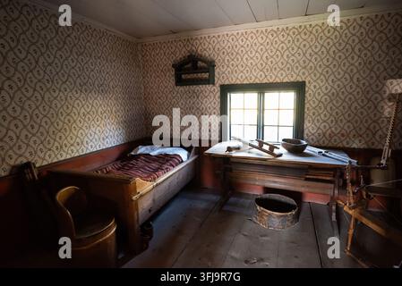 Rustikales historisches Schlafzimmer mit gemusterter Tapete, Holzbetten, Schreibtisch und Stuhl neben einem hellen Fenster in einem alten Landhaus. Stockfoto
