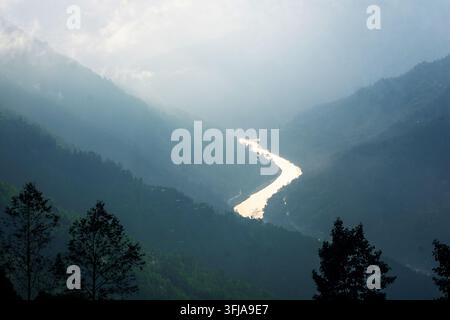 Ein ruhiger Fluss windet sich durch neblige Berge, die im sanften Sonnenlicht leuchten. Dunkle Silhouetten von Bäumen rahmen diese friedliche Landschaft ein. Stockfoto