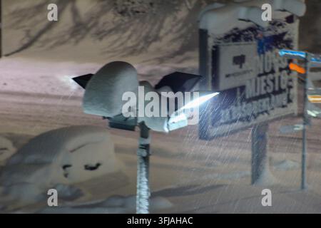 Eine stark mit Schnee bedeckte Ampel leuchtet durch fallende Schneeflocken in der Nacht, mit einem verschwommenen Ladenschild im Hintergrund. Stockfoto