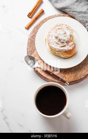 Frisch gebackener Zimtbrötchen mit weißer Zuckerglasur, Tasse schwarzer Kaffee und Gewürz auf einem hellen Marmorhintergrund. Ein gemütliches skandinavisches Frühstückskonzept fo Stockfoto
