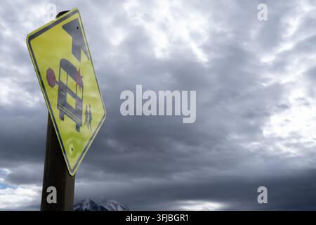 Gelbes Schild warnt vor Schulbus-Haltestelle voraus. Schild ist auf einem Holzpfosten montiert. Dunkle Wolken türmen sich über fernen Gipfeln Stockfoto