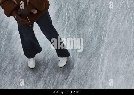 Eine Hochwinkel-Nahaufnahme einer Person, die weiße Eiskunstlaufschuhe trägt und auf einer zerkratzten Wintereisfläche gleitet Stockfoto